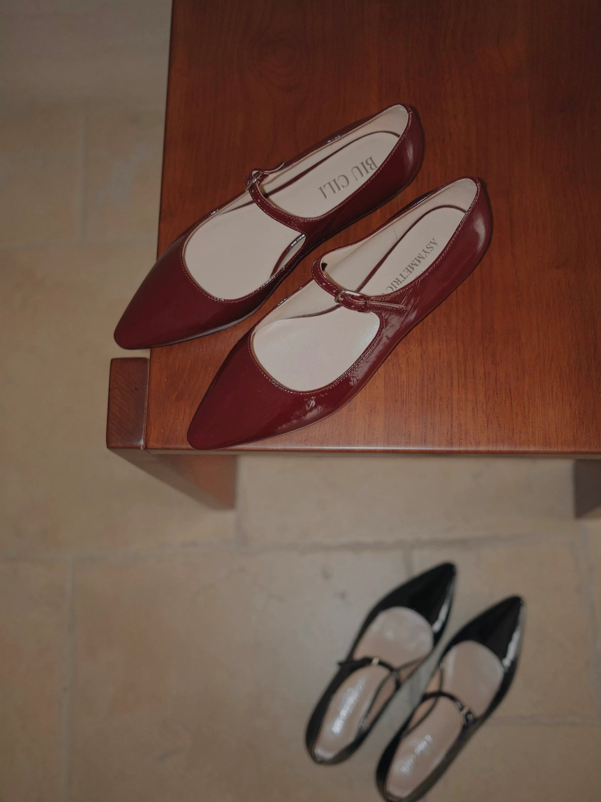 Red patent leather pointed-toe Mary Jane flats on a wooden surface, with a blurred black pair in the background