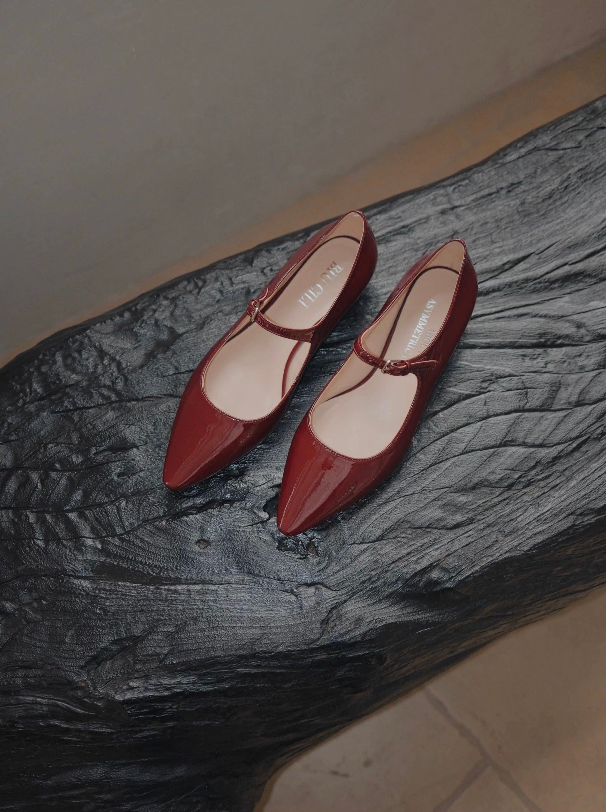 Pair of merlot red patent leather pointed-toe Mary Jane flats with a single strap, displayed on a sculptural black wooden surface