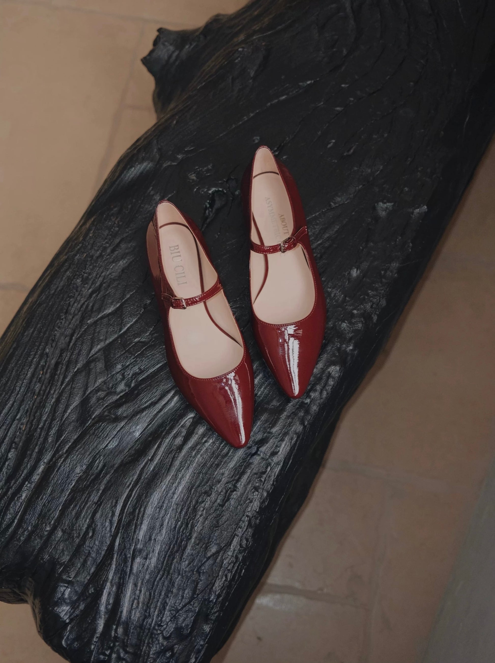 Pair of merlot red patent leather pointed-toe Mary Jane flats with a single strap, displayed on a sculptural black wooden surface