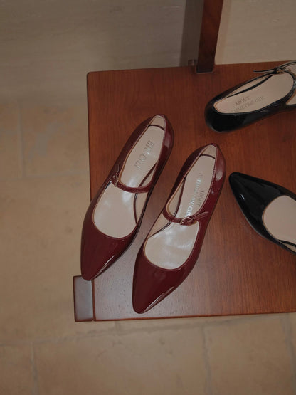 Red patent leather pointed-toe Mary Jane flats on a wooden surface, with a blurred black pair in the background