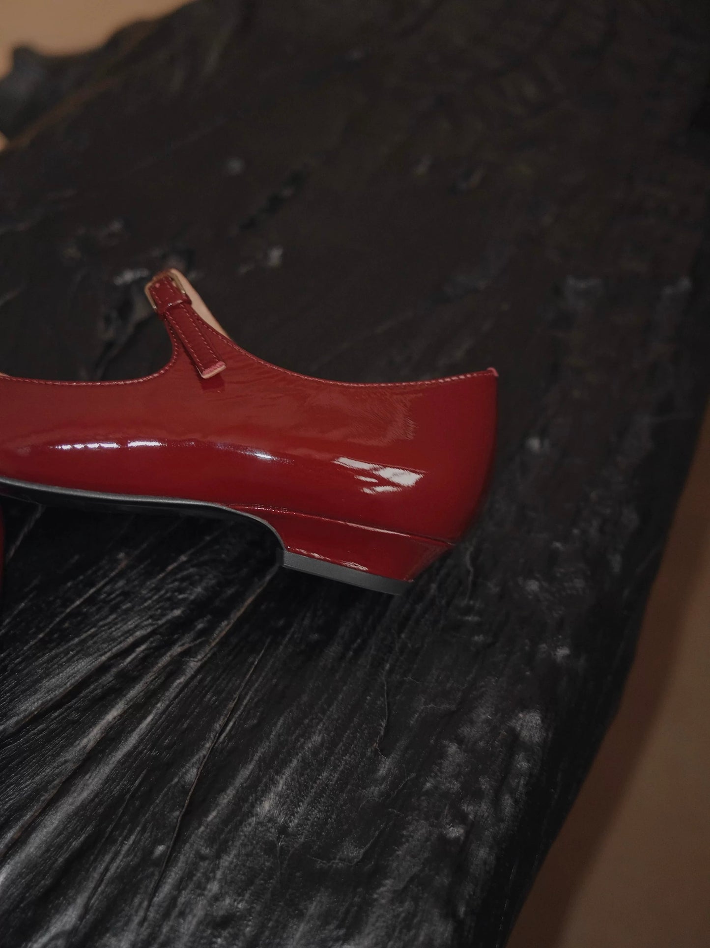 Showing the heel details of a deep red patent leather pointed-toe Mary Jane flats with a single strap, displayed on a sculptural black wooden surface
