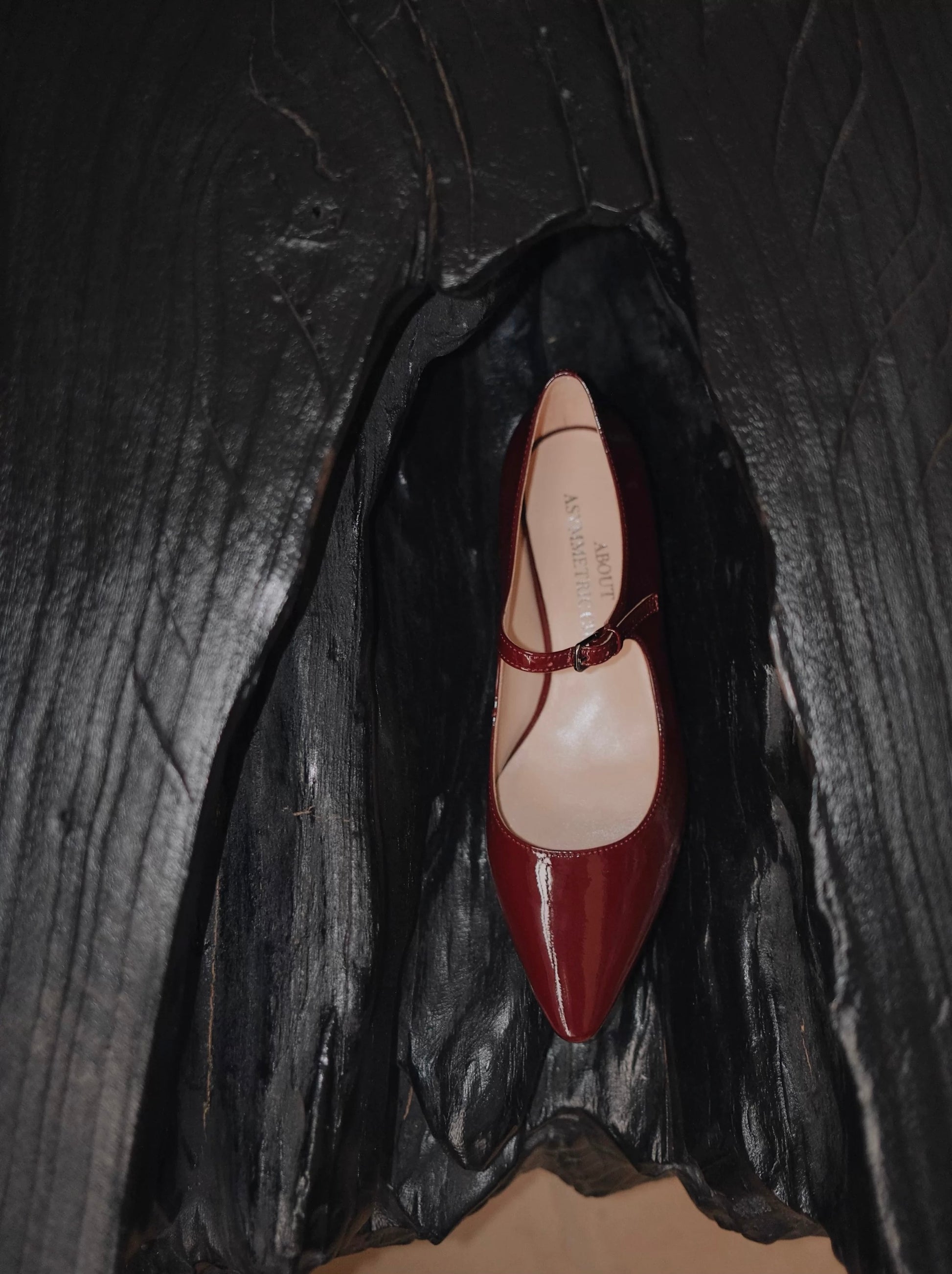 One deep red patent leather pointed-toe Mary Jane flats with a single strap, displayed on a sculptural black wooden surface