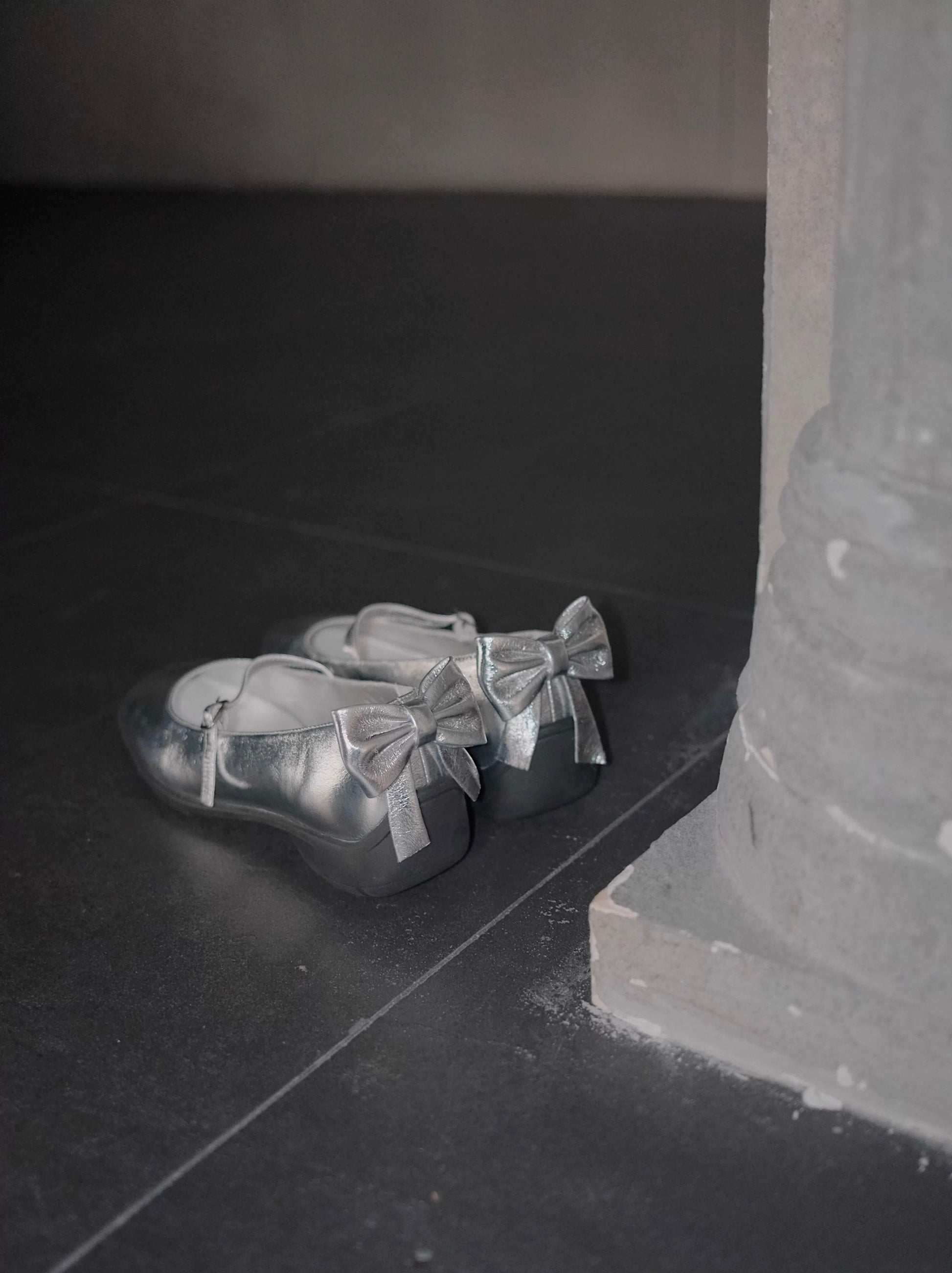 Sporty Mary Jane shoes in shimmering silver with back bow place on a dark rought floor - back view - showing the bow details