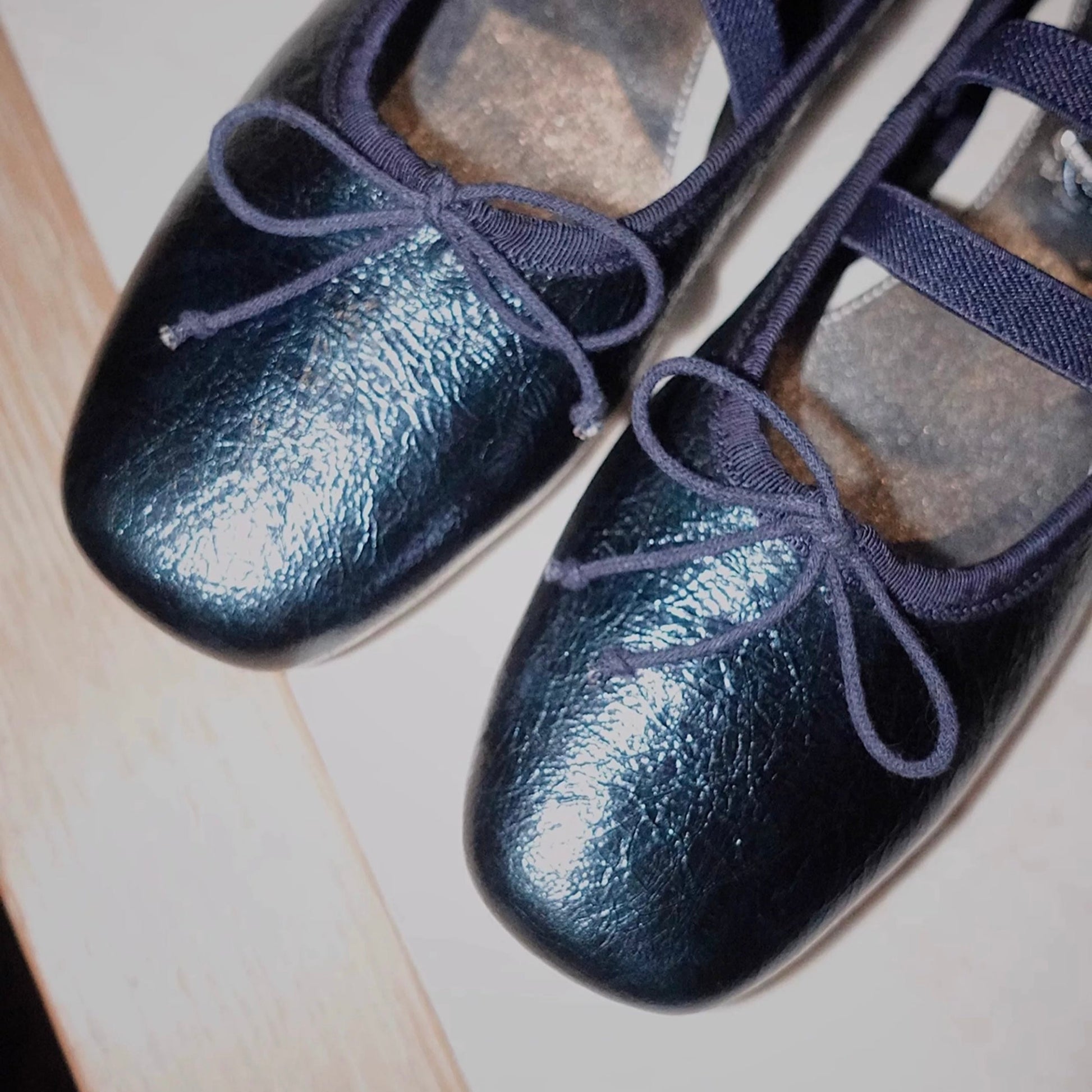 Closed view of the front of a pair of shimmering navy metallic leather Mary Jane ballet flats with asymmetrical cross straps