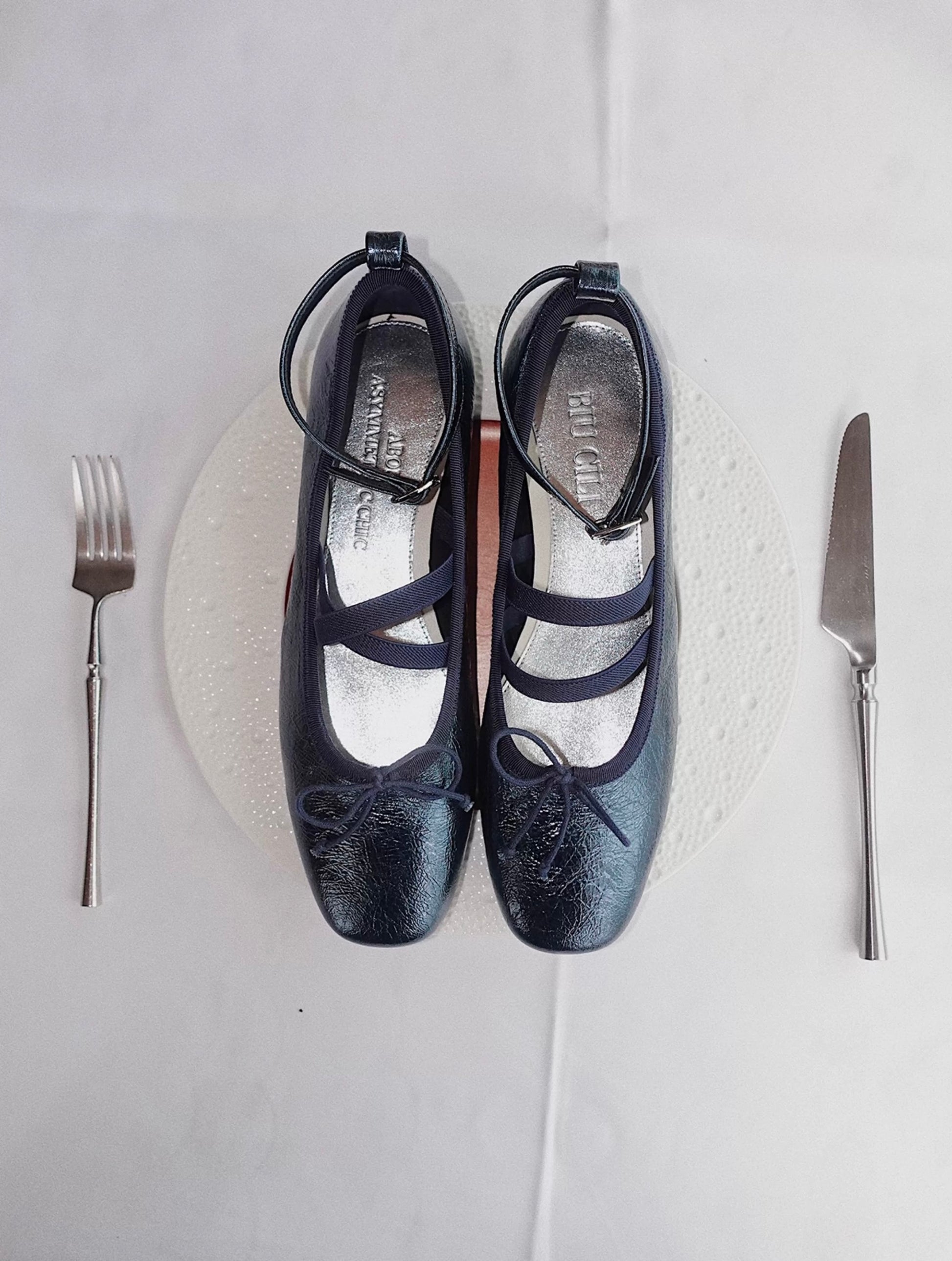 Navy metallic leather Mary Jane ballet flats with asymmetrical cross straps, styled playfully on a white plate with fork and knife as if served at a dinner setting