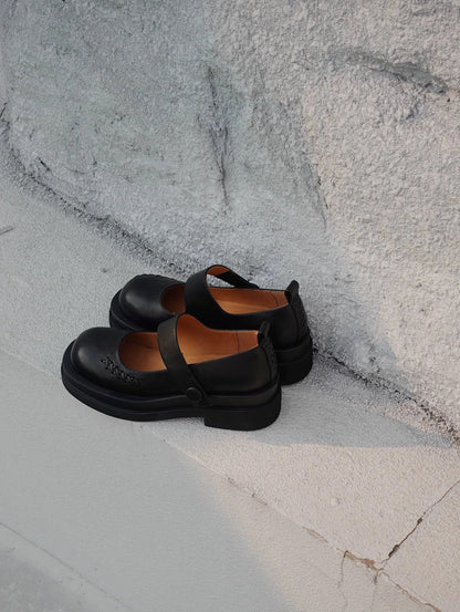 Black platform Mary Jane shoes with wide strap and low block heel, displayed on a textured concrete floor beside a wall