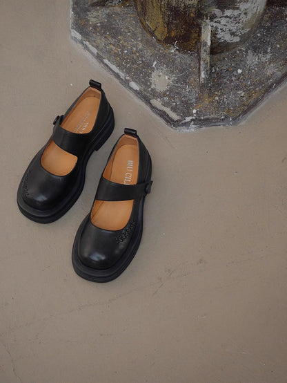 Black platform Mary Jane shoes with wide strap and low block heel, displayed on a textured concrete floor beside a wall