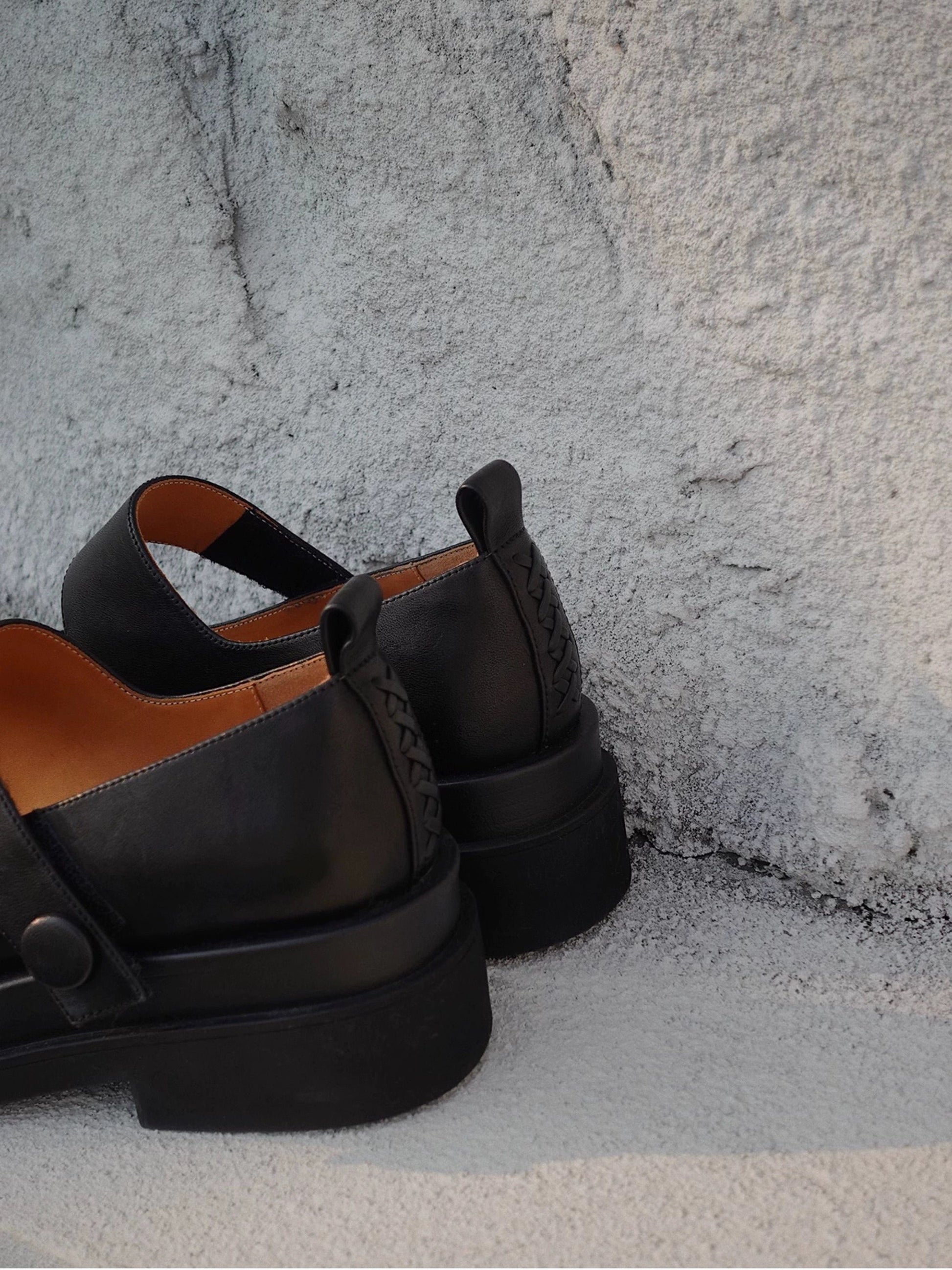Black platform Mary Jane shoes with wide strap and low block heel, made with premium horsehide leather, displayed on a textured concrete floor beside a wall