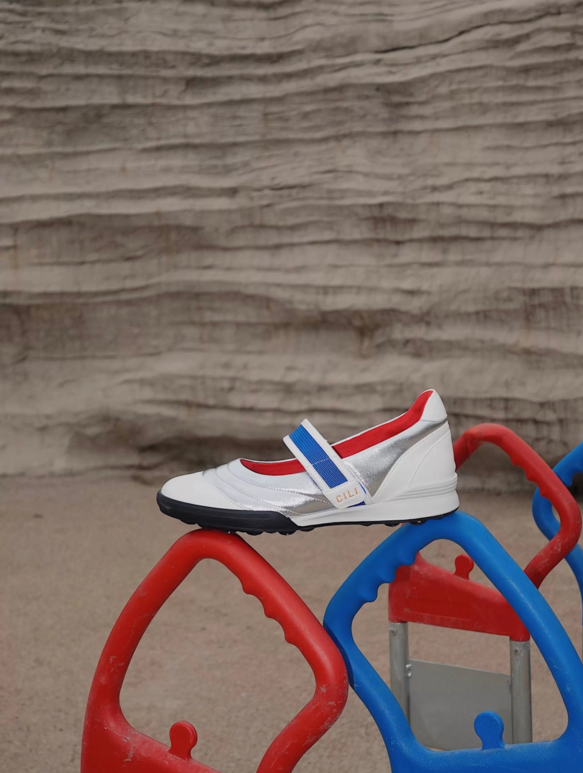 Kaia asymmetric Mary Jane flats in silver, white leather, red lining, with a blue strap, styled on colorful playground bars for a sporty-chic look