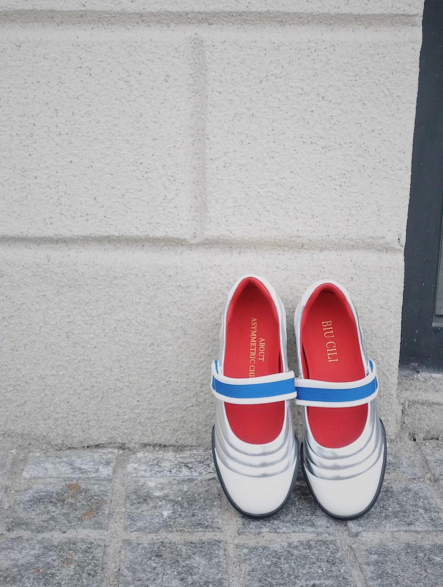 Pair of Kaia leather Mary Jane flats in colorblock white and silver with red lining and blue strap, positioned against a white textured wall