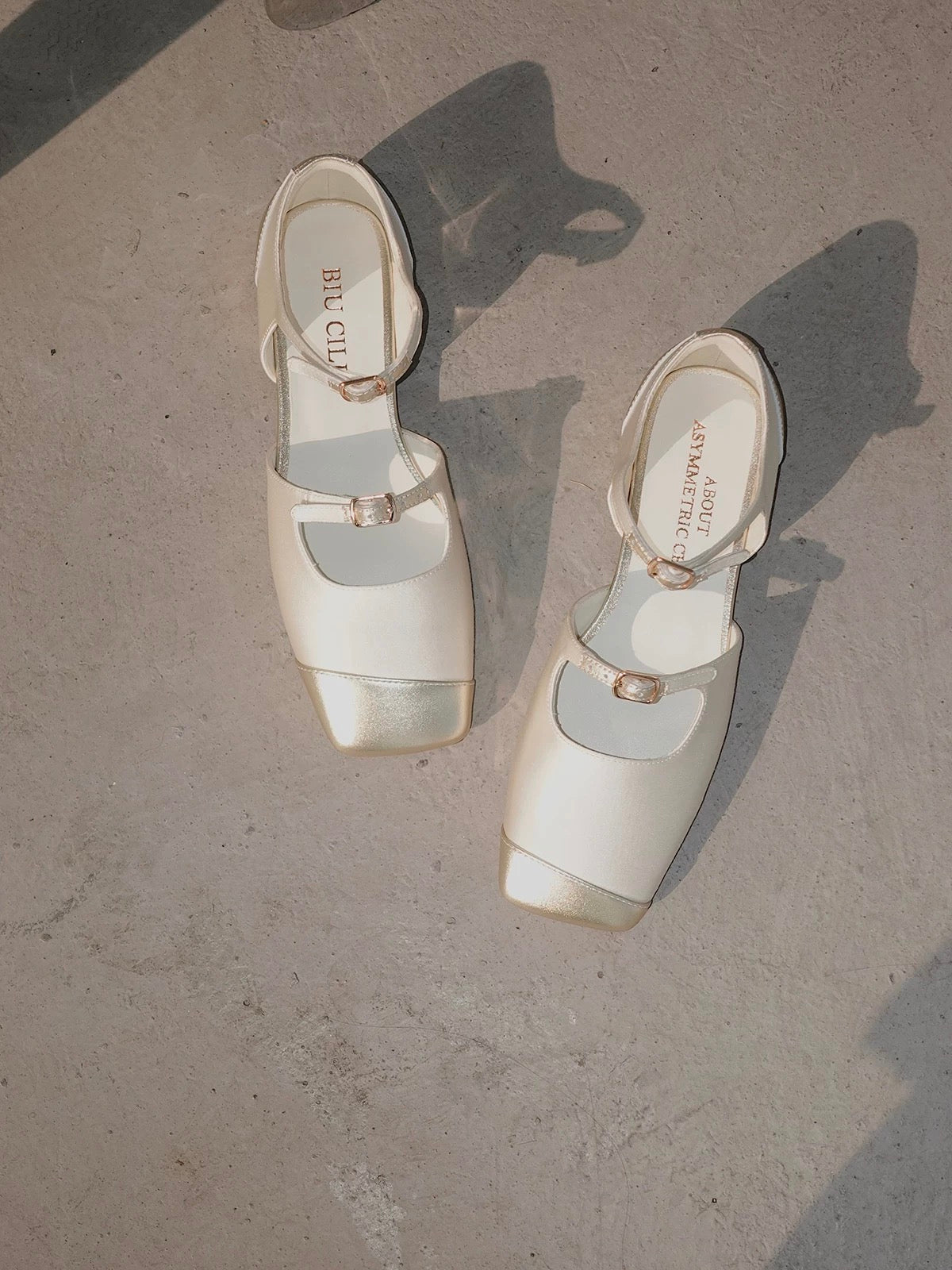 A pair of white silk mary jane flats with straps and asymmetric gold leather details at toe front, placed on the floor