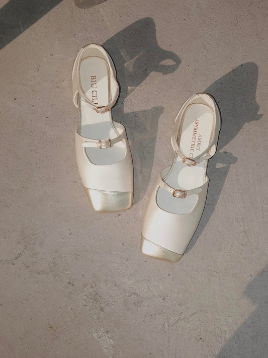 A pair of white silk mary jane flats with straps and asymmetric gold leather details at toe front, placed on the floor