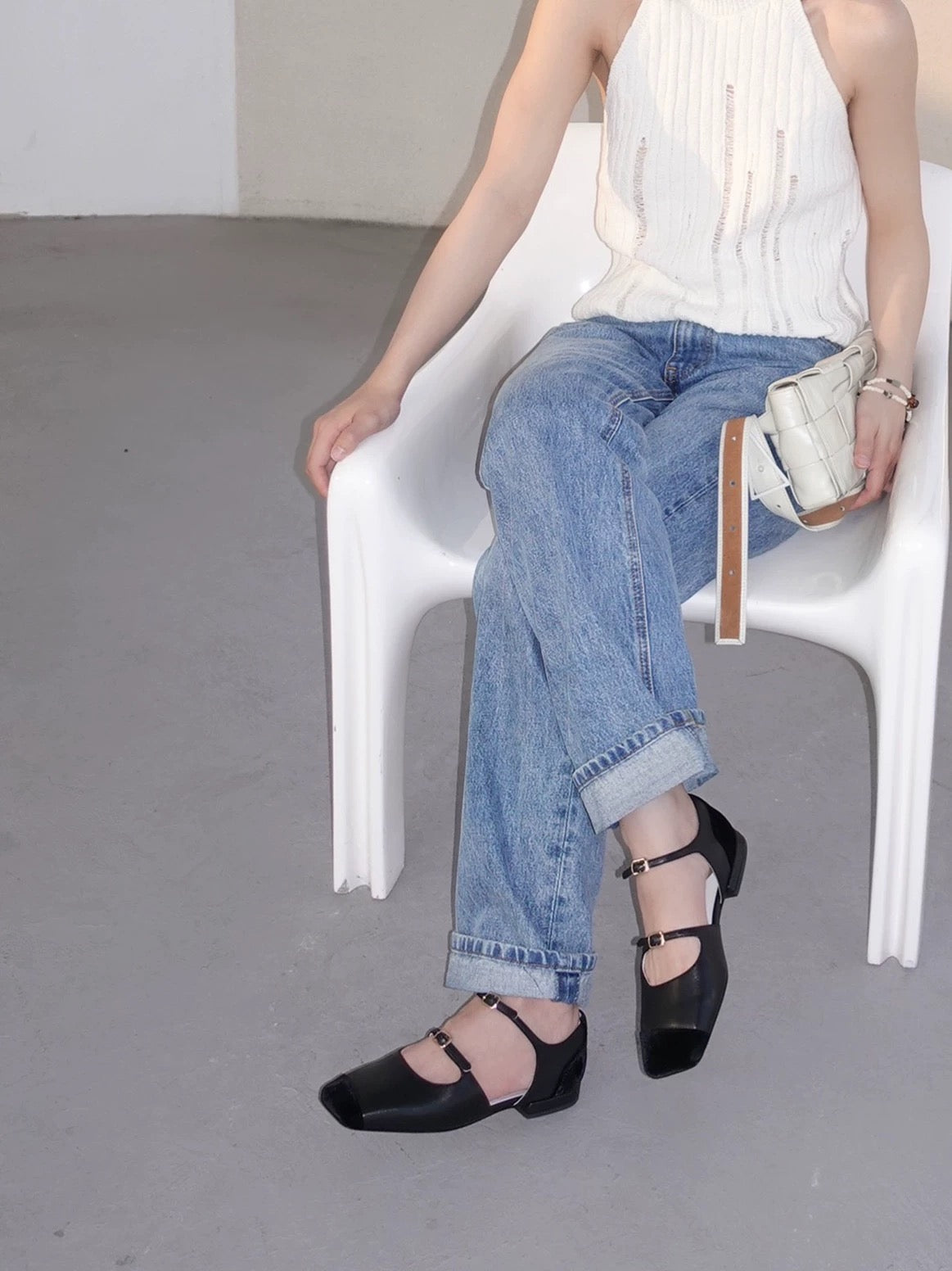 Women wearing a pair of black leather mary jane flats with straps and asymmetric velvet details at toe front, metallic buckles, pairing with jeans and white top, sitting on a white chair