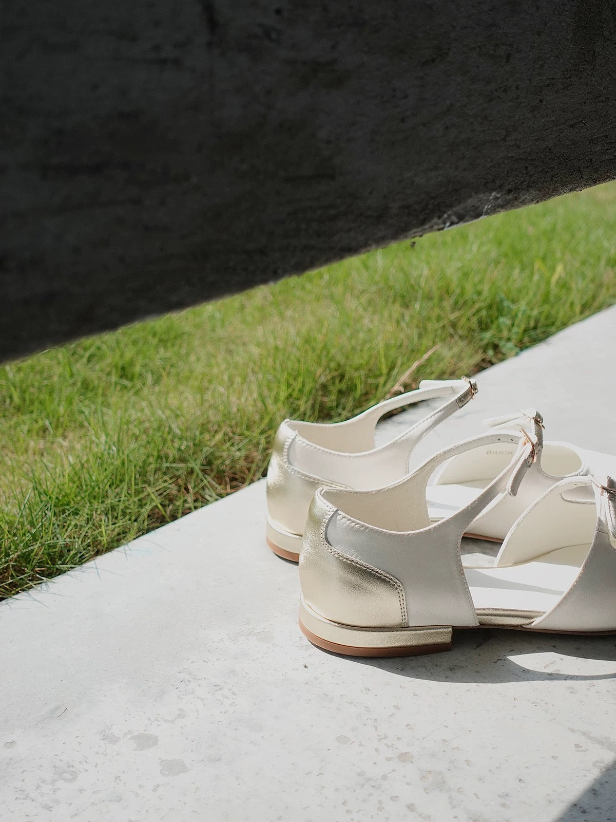 Closed back view of the gold asymmetric leather front of a pair of white silk mary jane flats with straps, showing the gold details