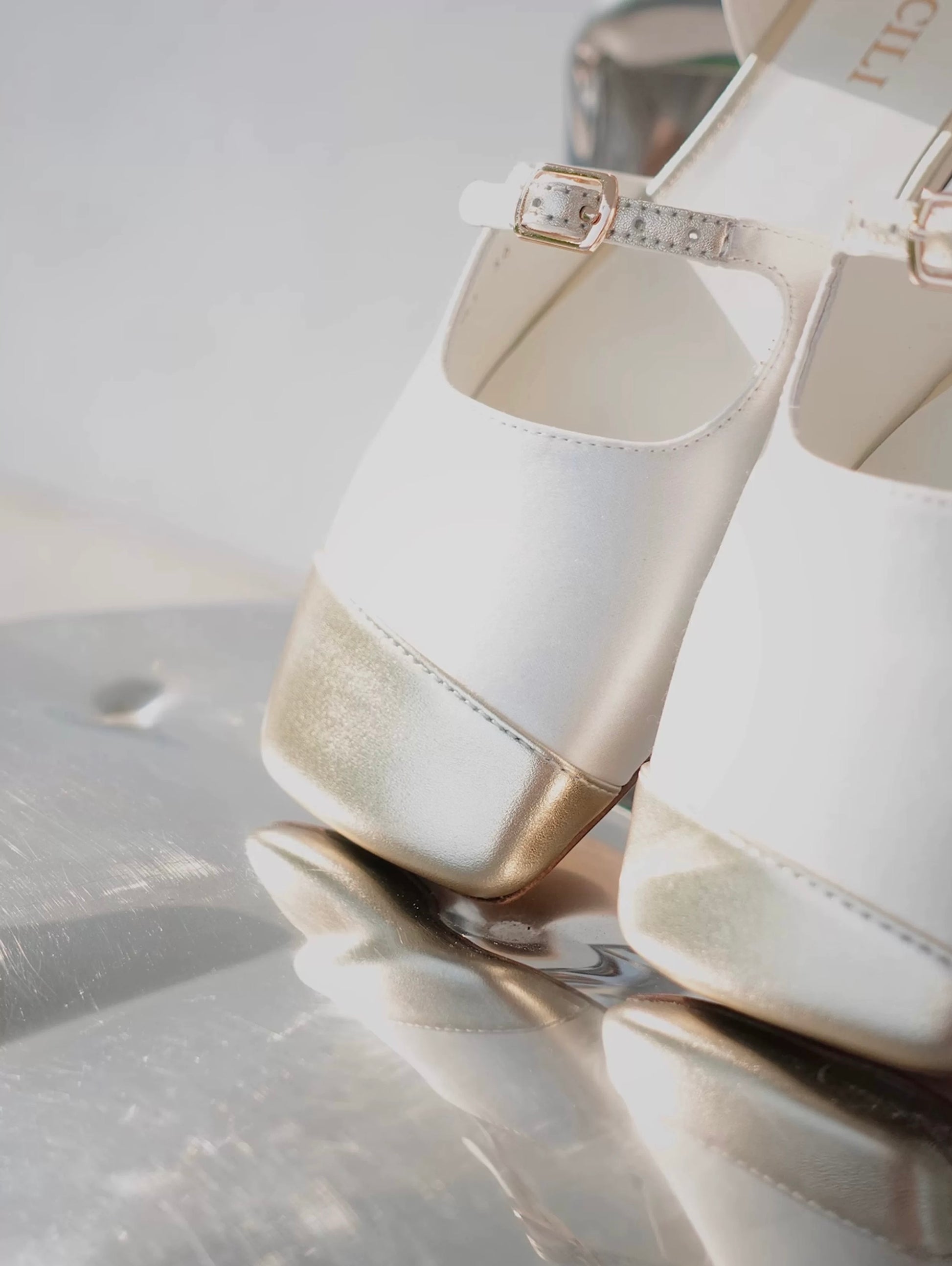 Closed view of the gold asymmetric leather front of a pair of white silk mary jane flats with straps