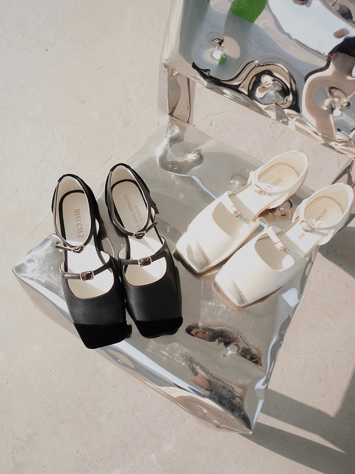 A pair of white silk mary jane flats with straps and asymmetric gold leather details at toe front, and a pair of black leather mary jane flats with straps and asymmetric velvet details at toe front, placed on a shiny mirror like chair