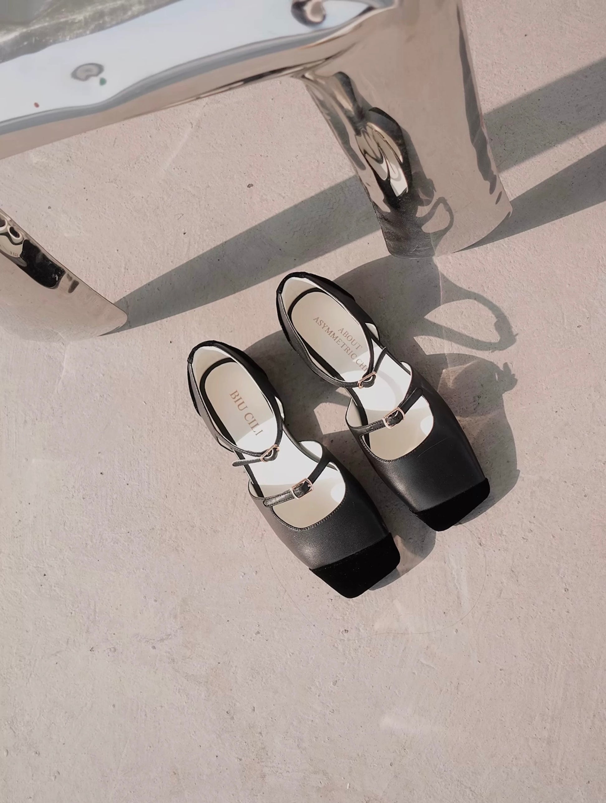 Top view of a pair of black leather mary jane flats with straps and asymmetric velvet details at toe front, placed on the floor next to a chair