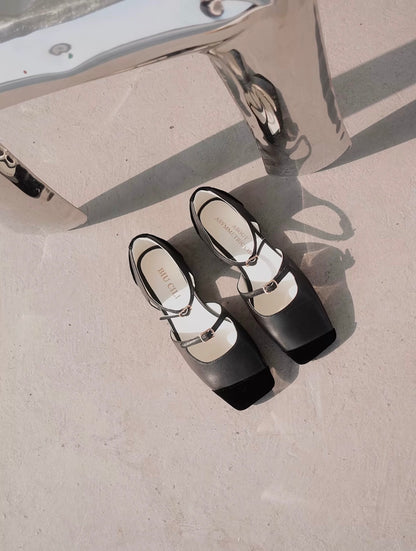 Top view of a pair of black leather mary jane flats with straps and asymmetric velvet details at toe front, placed on the floor next to a chair