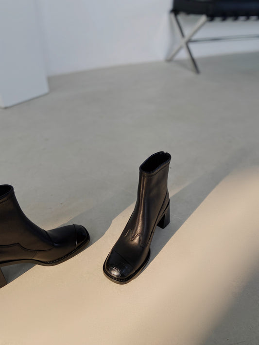 Women's black leather square-toe ankle boots with block heels, styled against a concrete wall in warm natural light