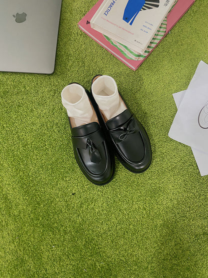 Black leather loafers for women featuring asymmetric tie and bow details, crafted in matte calfskin with low heel and soft structure, place on grass.
