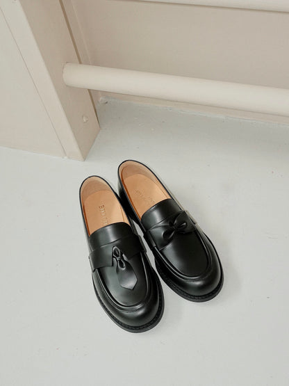 Black leather loafers for women featuring asymmetric tie and bow details, crafted in matte calfskin with low heel and soft structure, placed on the ground