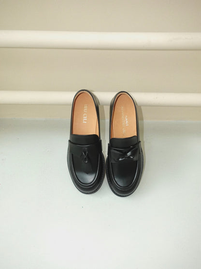 Black leather loafers for women featuring asymmetric tie and bow details, crafted in matte calfskin with low heel and soft structure, placed on the ground.