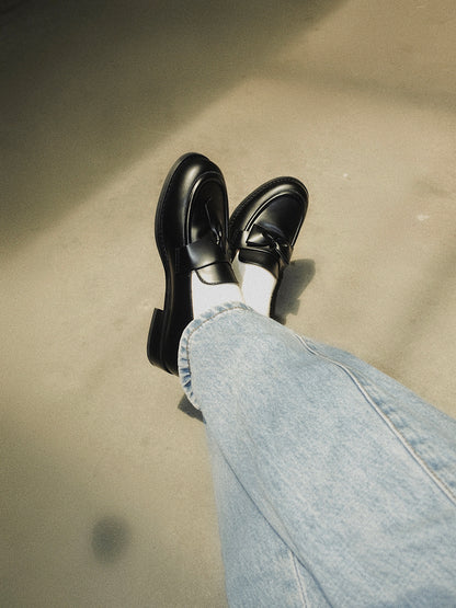Women wearing a pair of black leather loafers featuring asymmetric tie and bow details, crafted in matte calfskin with low heel and soft structure, sitting on the floor