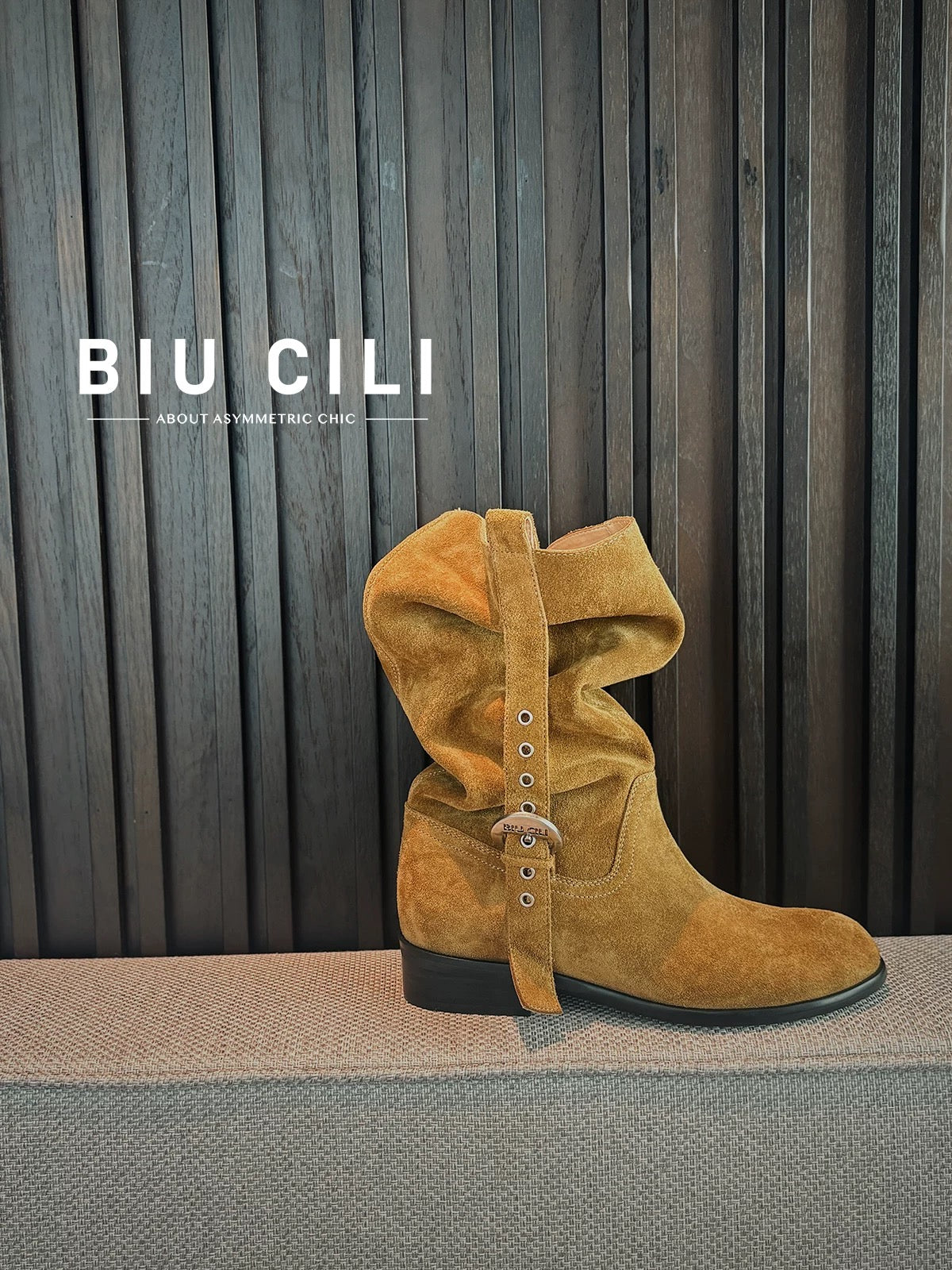 Brown suede boot with decorative straps on a gray surface against a striped curtain background, featuring the brand 'BIU CILI'.