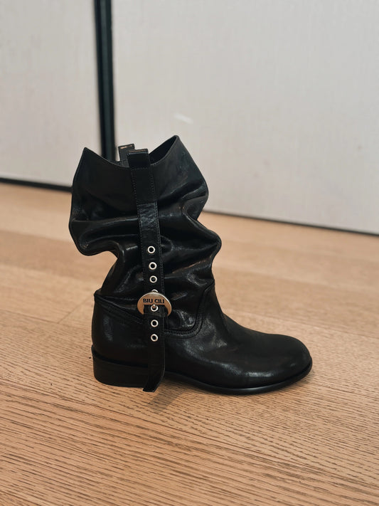 Black leather boot with decorative straps on a wooden floor.