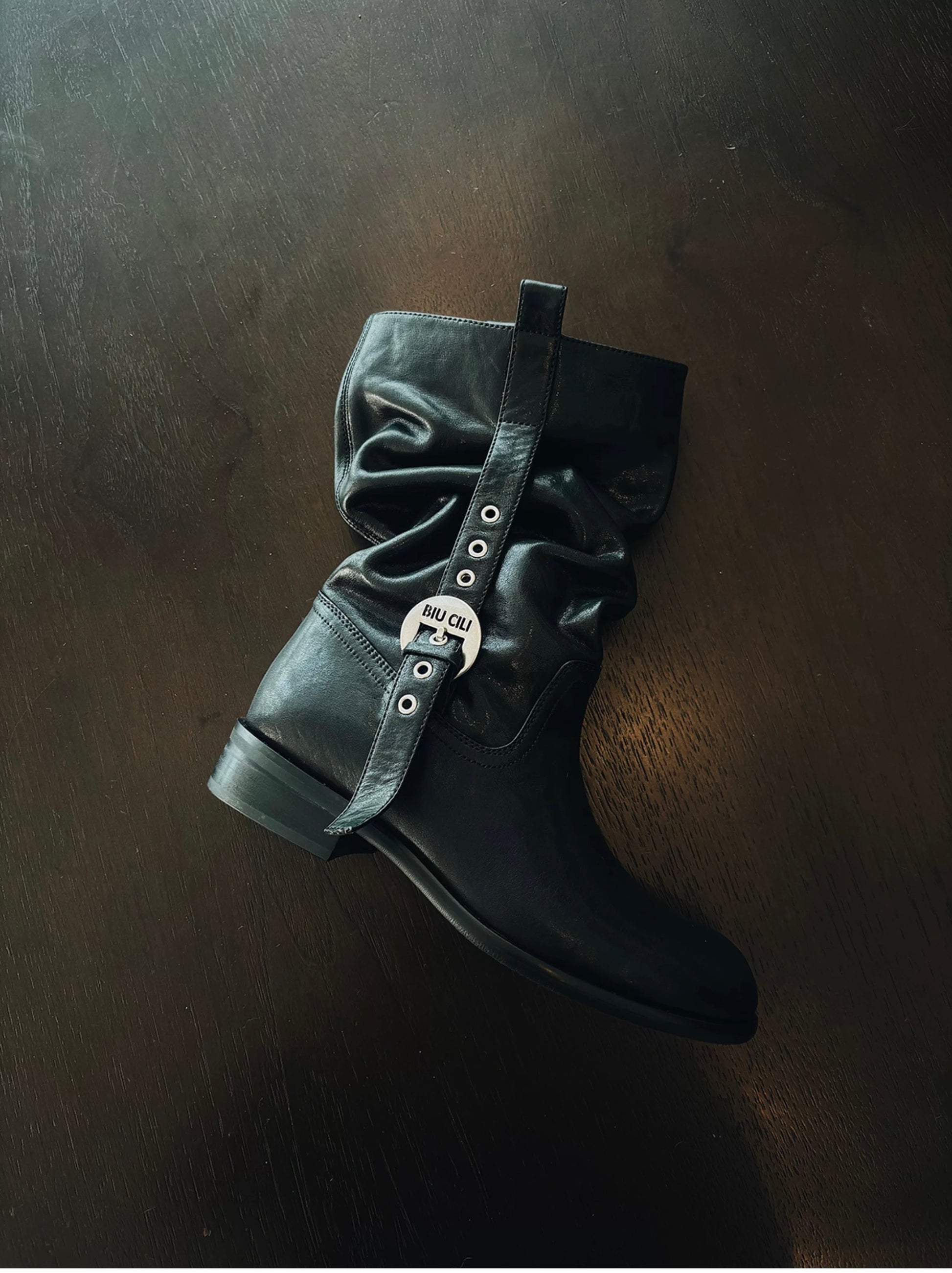 Black mid-calf leather boot with decorative buckle on a dark surface
