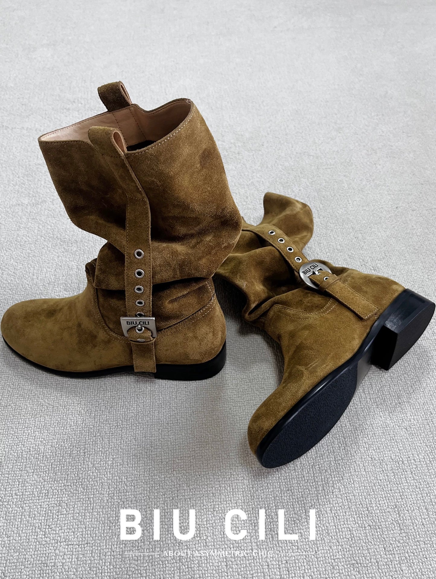 Brown suede boots with asymmetric metal buckles on a light gray background, featuring the brand 'BIU CILI'.