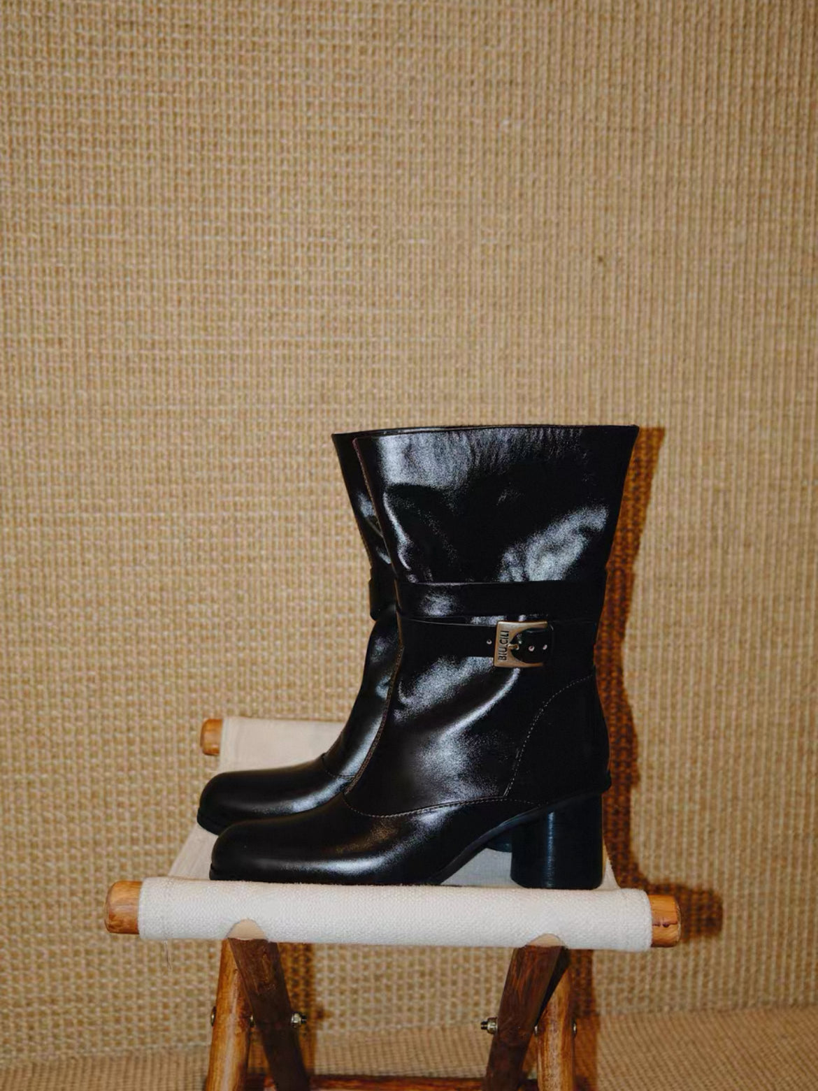 Black leather boot on a small wooden stool against a textured beige wall.