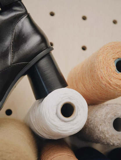 The block heel detail  of black leather boot being used to unwind white yarn from a spool on a pegboard.