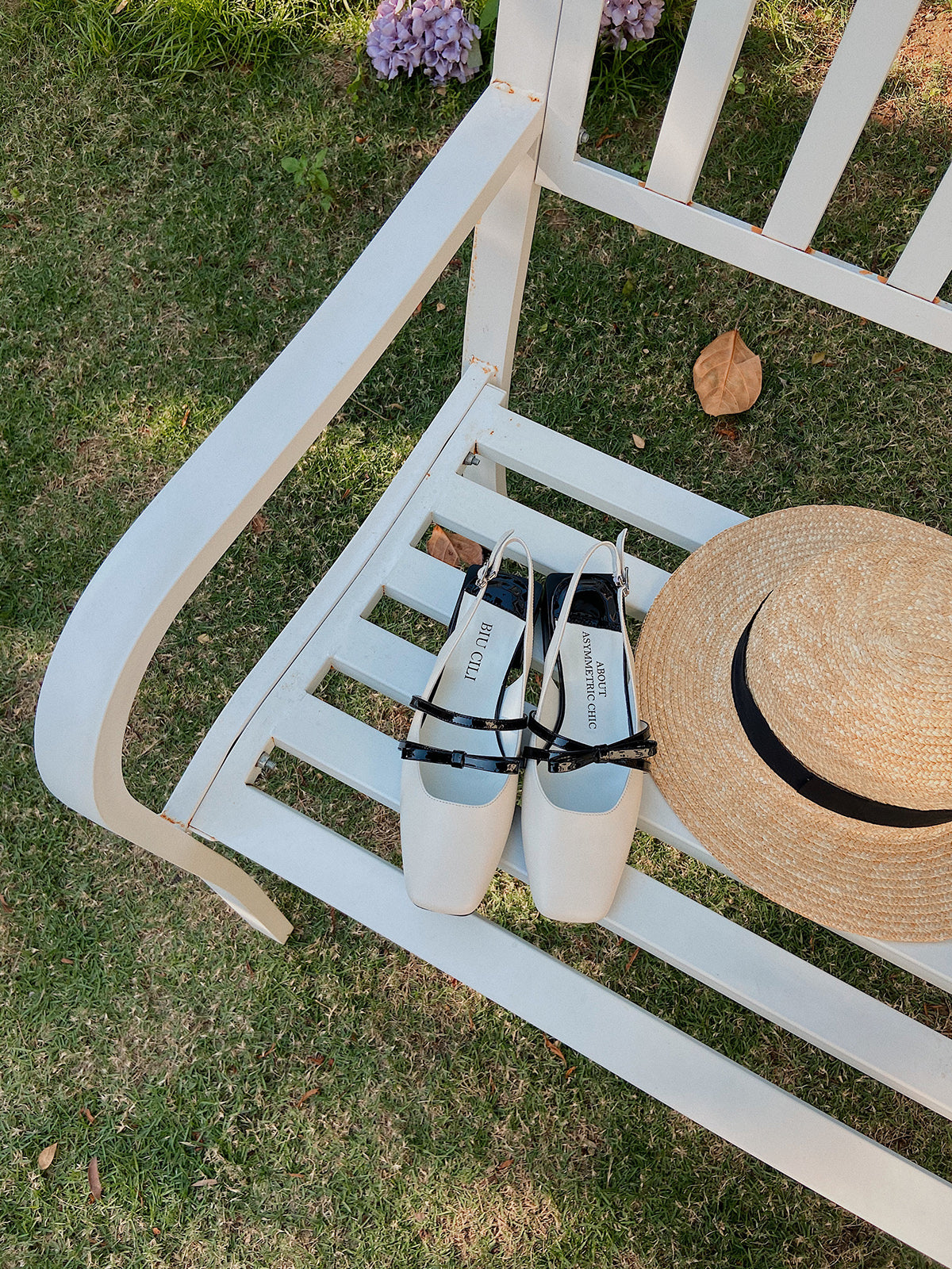 White Mary Jane shoes with square toe and black asymmetric bow straps, contrast heel, and soft leather finish, near a hat
