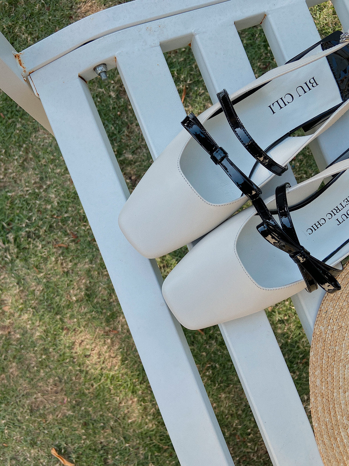 White Mary Jane shoes with square toe and black asymmetric bow straps, contrast heel, and soft leather finish, placed on a bench