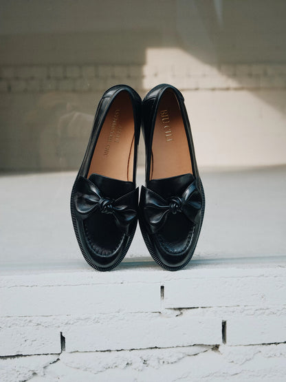 Pair of black loafers with bow details on a white wooden floor.