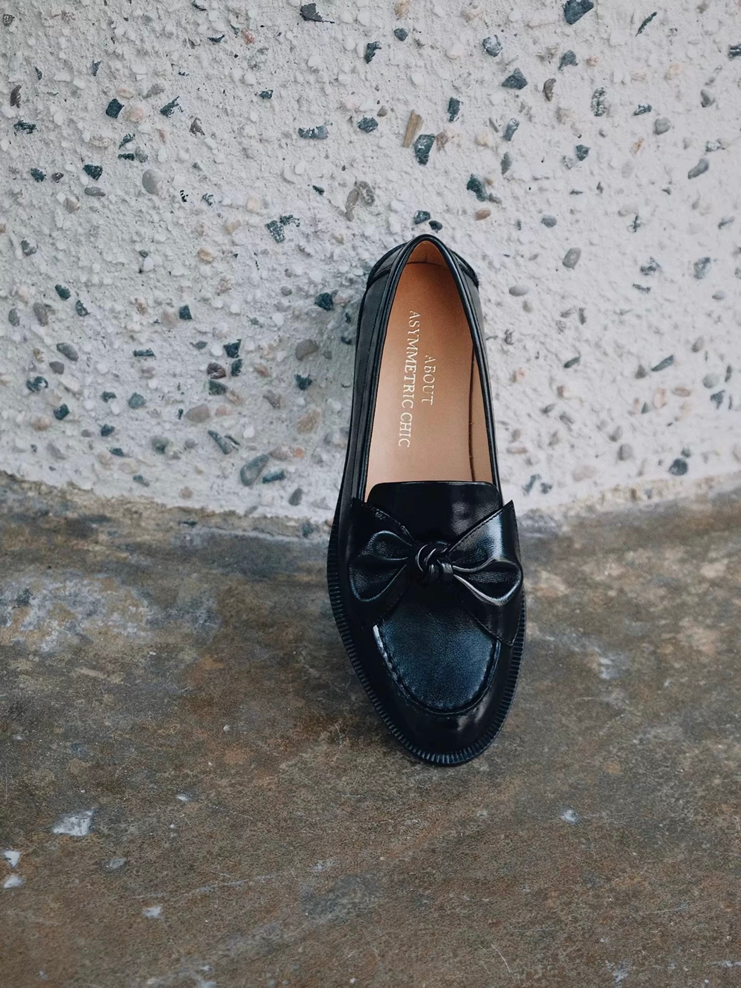 Black loafer shoe with a bow on a textured concrete surface