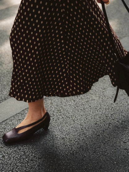 Person wearing a black polka dot dress and plum brown bock heel shoes with cut-out on a pavement.