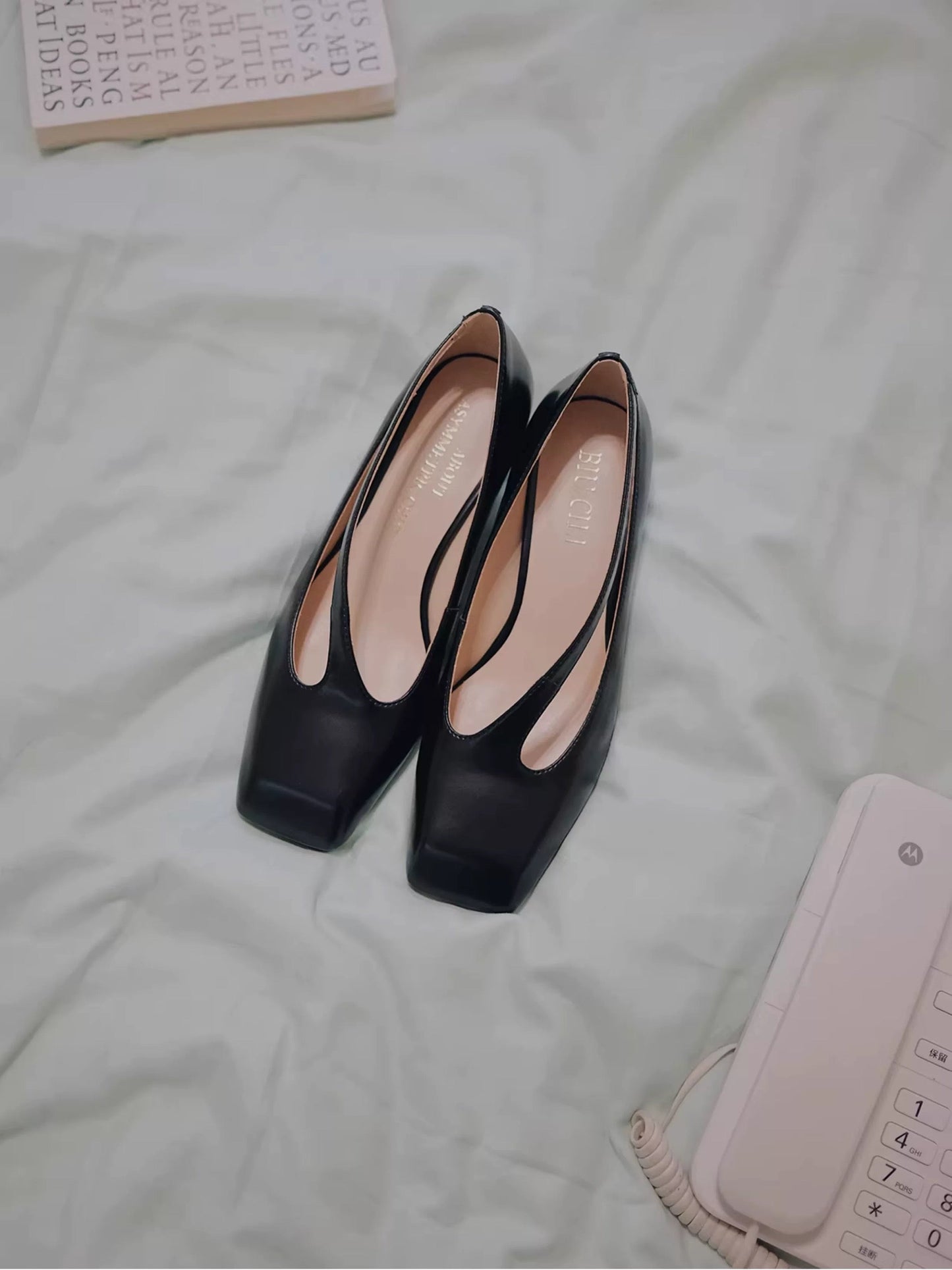 Black high-heeled shoes on a light-colored surface with a phone and book in the background.