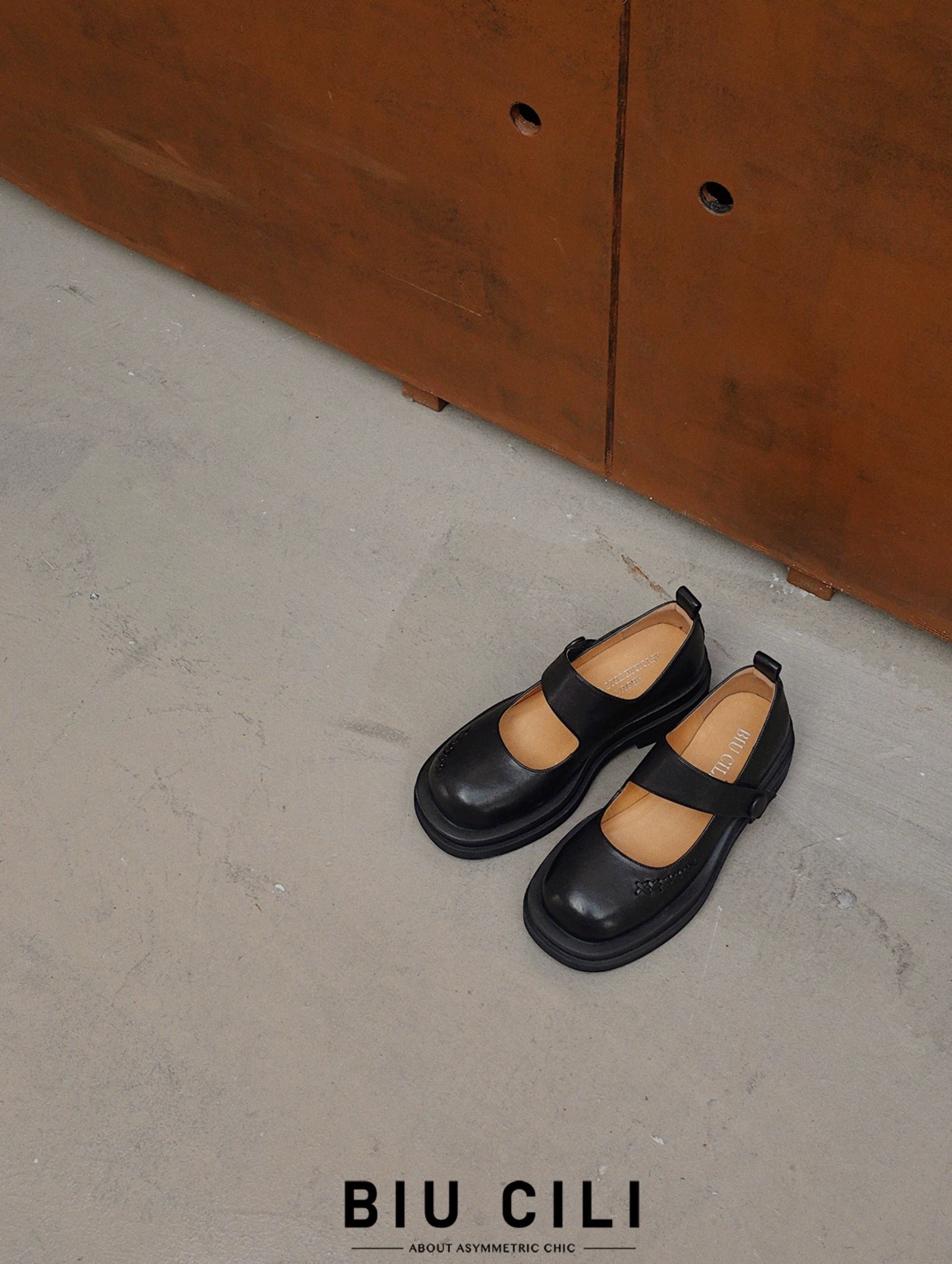 Black platform Mary Jane shoes with wide strap and low block heel, displayed on a textured concrete floor beside a rust-toned cabinet