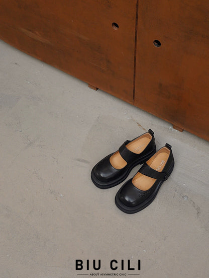 Black platform Mary Jane shoes with wide strap and low block heel, displayed on a textured concrete floor beside a rust-toned cabinet